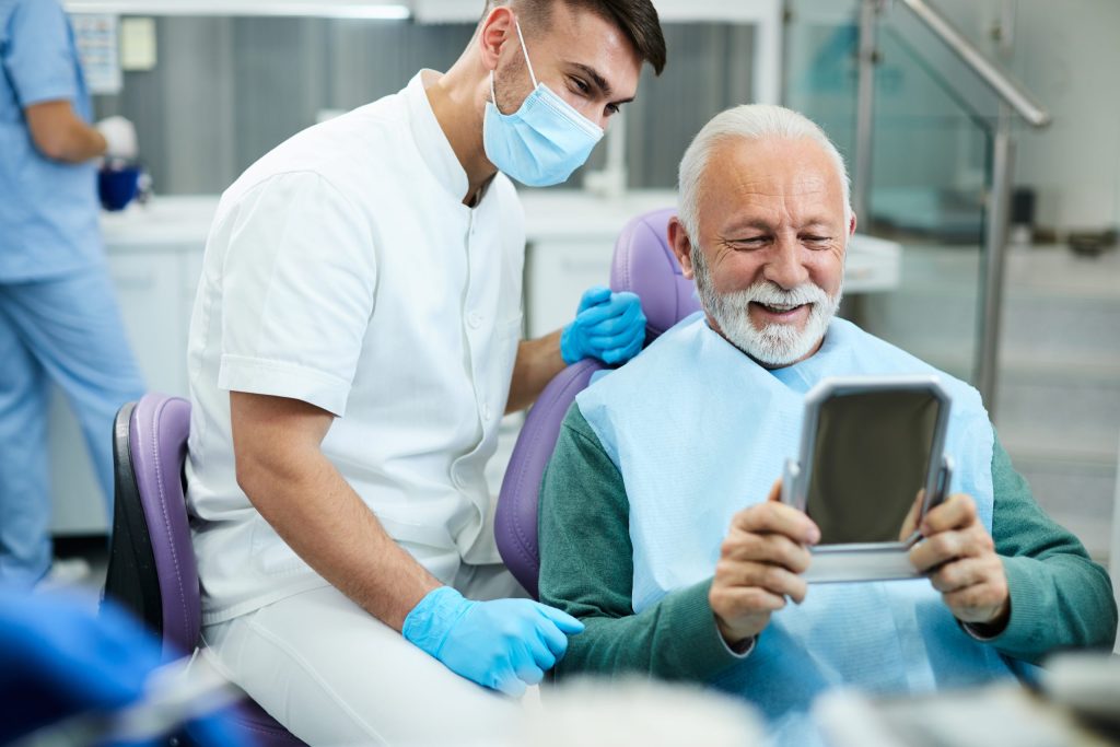 Smiling senior patient checking his teeth in a hand mirror beside his dentist