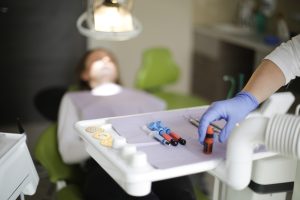 Blurred patient reclining in a dental chair while a gloved hand arranges syringes and vials on an instrument tray