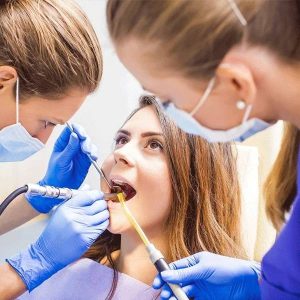 Dental professionals in blue gloves examine a patient's teeth with clinical instruments in a bright dental office
